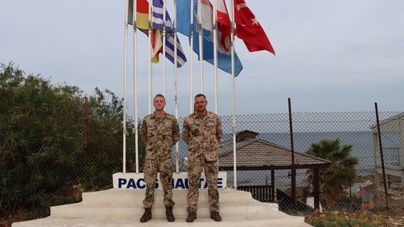 Zwei Soldaten stehen in sandfarbenen Uniformen vor mehreren Fahnenmasten © Bundeswehr 