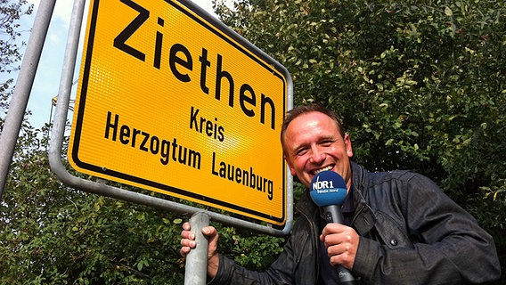 Moderator Jan Bastick vor dem Ortsschild Ziehten mit einem Mikrofon in der Hand Moderator Jan Bastick vor dem Ortsschild Ziehten mit einem Mikrofon in der Hand © NDR Foto: Karin Hauschildt