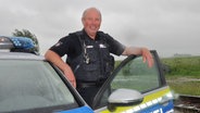 Polizist Bernd Busch steht in der offenen Fahrertür seines Polizeiautos. © NDR Foto: Peter Bartelt