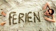 Ein Junge hat Sand auf seinem Körper und lächelt in die Kamera. Auf dem Sand ist der Schriftzug "Ferien" zu erkennen.  Foto: Jens Büttner