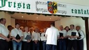 Ein Männerchor steht unter einem Banner mit dem Wappen der Friesen darauf. © NDR Foto: Karin Haug