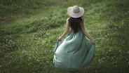 Eine Frau im grünen Kleid und Hut auf einer Wiese von hinten aufgenommen. © picture alliance Bildagentur-online Images Foto: Valeriya Tikho
