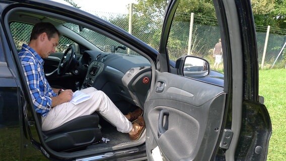 Meeno Schrader sitzt lesend im Auto bei geöffneter Tür © NDR Foto: Juliane Thomas
