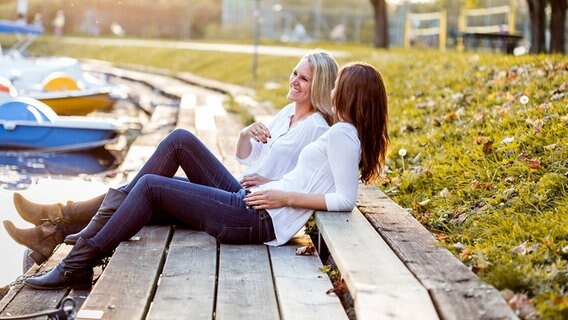 Zwei Frauen sitzen auf dem Steg an einem Paddelteich und lachen, tragen Jeans und Stiefel. © Imago/westend61 Foto: westend61