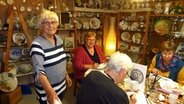 Drei ältere Frauen sitzen in einem Raum voller Porzellan und malen auf Porzellan und eine andere Frau steht daneben. © NDR Foto: Peer-Axel Kroeske