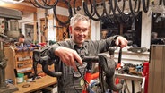 Ein Mann steht mit den Händen auf einem Fahrrad in einer Werkstatt. © NDR Foto: Jönr Schaar