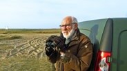 Peter Hering steht neben einem Auto an der Küste und hält sein Fernglas vor sich in der Hand und ist bereit hindurch zu spähen. © NDR Foto: Peer-Axel Kroeske