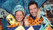 Nicole Bäumer und Martin Dronsfield stehen verkleidet unter einem bunten Schirm und lachen in die Kamera. © NDR Foto: Robert Tschuschke