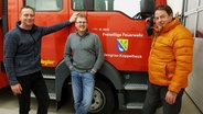 Volker Jürgensen, Lars Neumann und Björn Rohr vor einem Fahrzeug der Freiwilligen Feuerwehr Niesgrau. © NDR Foto: Lukas Knauer