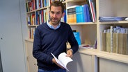 Skandinavist Prof. Steffen Höder lehnt sich lässig an ein Bücherregal und blättert in einem Buch. © NDR Foto: Andrea Ring