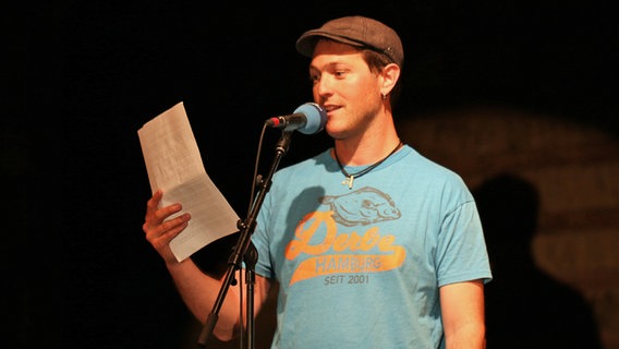 Geritt Hoss spricht auf einer Bühne in ein Mikrofon beim Poetry Slam op Platt in der Winkelscheune in Molfsee. © NDR Foto: Lornz Lorenzen