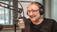 Der Schauspieler Rainer Bock sitzt vor einem Mikrofon in einem Hörfunkstudio. © NDR Foto: Janis Röhlig