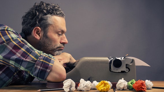 Ein Mann schaut frustriert auf eine Schreibmaschine. Im Vordergrund befinden sich zerknüllte Papiere. © photocase.de Foto: Andreas Berheide