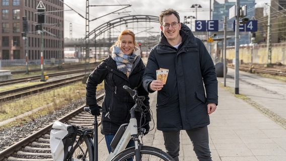 Die NDR-Moderatoren Mandy Schmidt (links) und Horst Hoof (rechts) stehen mit einem E-Fahrrad auf einem Bahnsteig und halten zwei 50-Euro-Scheine in die Kamera. © NDR Foto: Janis Röhlig