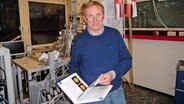 Der Physiker Holger Kersten steht mit einem Buch in der Hand in einer Werkstatt. © Andrea Ring Foto: Andrea Ring