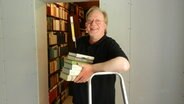 Ein Mann steht vor einer geöffneten Tür zu einem Raum voller gefüllter Bücherregale. Auf seinem Arm hält er einen Stapel Bücher. © Ines Barber Foto: Ines Barber