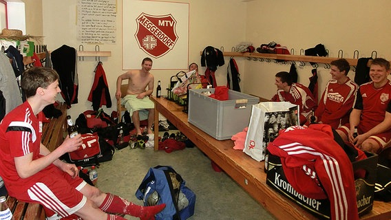 Sechs Spieler sitzen in einer Fußballkabine, an der Wand prangt das Logo des MTV Meggerdorf. © NDR Foto: Dana Frohbös