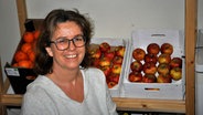 Claudia Laparose sitzt vor einem Regal, in dem 3 Kisten mit Äpfeln stehen und schaut lächelnd in die Kamera. © NDR Foto: Frank Hajasch
