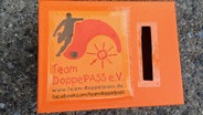 Eine orangefarbene Box mit der Aufschrift Team DoppelPASS e.V. und einem Schlitz © NDR Foto: Iris Guhl