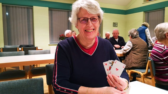 Rentnerin Karin Kühlmann sitzt in einem Raum voller Senioren und hält lächelnd Spielkarten in einer Hand © NDR Foto: Simone Steinhardt