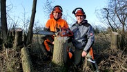 Dirk Lafrenz und Lehrgangsdeelnehmer Jan Kiehne sitzen mit ihrer Kettensäge vor einem Baumstumpf. © NDR Foto: Dana Frohbös