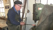 Der Schmied steht vor einem Federhammer und benutzt ihn zum bearbeiten seiner glühenden Eisenstange. © NDR Foto: Dana Frohbös