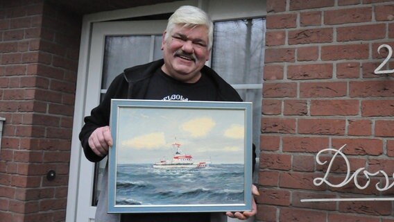 Ein Mann lächelt in die Kamera und hält ein Gemälde hoch, auf dem ein Seenotrettungsschiff zu sehen ist. © NDR Foto: Peter Bartelt