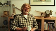 Arved Fuchs sitzt mit einer Kaffeetasse in der Hand vor seinem Bücherregal und lächelt in die Kamera. © NDR Foto: Doreen Pelz