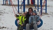 Eine Frau und zwei kleine Kinder sitzen auf einer Schaukel auf einem Spielplatz. © NDR Foto: Nadine Dietrich