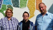 Drei Personen stehen vor einem Mosaikbild. Von Links nach Rechts stehen Sabine Graf, ihr Sohn Christoph und der Vater Andreas Volkmar vor dem Bild. © NDR Foto: Frank Hajasch