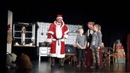 Ein als Nikolaus verkleideter Mann steht auf der Bühne. Daneben schauen sich ein Junge und ein Mädchen an, während eine Frau in ihrer Mitte auf sie herab blickt. © NDR Foto: Karin Haug