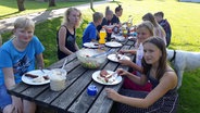 Zahlreiche Jugendliche sitzen bei einem gemütlichen Essen an einem Picknicktisch. © NDR Foto: Karin Haug