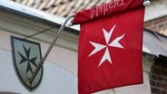 Eine Flagge mit dem rot-weißem Logo der Malteser hängt seitlich an einer Außenwand. © imago images Foto: Manngold