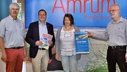 Drei Männer und eine Frau stehen vor einem Aufsteller mit der Aufschrift "Amrum" und halten lächelnd Prospekte in die Kamera. © Nordfriisk Instituut Foto: Harry Kunz