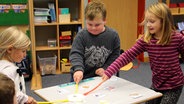 Drei Kinder stehen um einen kleinen Tisch und hauen mit einer Fliegenklatsche auf Bilder. © NDR Foto: Rebekka Merholz