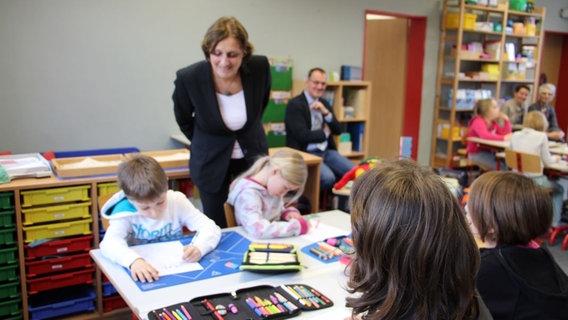 Fünf Kinder sitzen an einem Gruppentisch während des Plattdeutsch Unterrichts. Im Hintergrund sitzen mehrere Erwachsene und beobachten den Unterricht. © NDR Foto: Rebekka Merholz