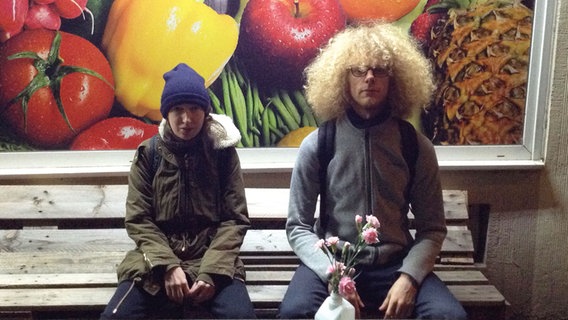 Die Band Tüdelband sitzt in winterlicher Kleidung auf einer Bank, im Hintergrund ist ein Plakat mit Obst und Gemüse. © Mire Buthmann Foto: Mire Buthmann