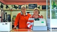 Angelika Micheel und Elke Wandel stehen hinter der Kasse, im Hintergrund sind beschriftete Tafeln zu sehen. © NDR Foto: Andrea Ring