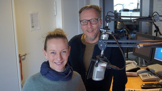Katja Just und Jan Malte Andresen stehen im NDR 1 Welle Nord Sendestudio neben zwei Mikrofonen und lächeln in die Kamera. © NDR Foto: Samir Chawki