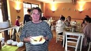 Susanne Lühr steht in ihrem Cafe mit einer Schüssel in der Hand. Im Hintergrund sitzen Gäste an Tischen. © NDR Foto: Peer-Axel Kroeske