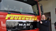 Daniel Schultz steht vor einem Feuerwehrfahrzeug und zeigt mit beiden Händen auf den Schriftzug, der auf dem Auto ist. © NDR Foto: Jörn Schaar