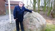 Birger Feder steht in einer Feuerwehruniform vor einem großen Szein, auf dem "Freiwillige Feuerwehr Löwenstedt" eingemeißelt ist und legt seine Hand auf diesen Stein. © NDR Foto: Peer-Axel Kroeske