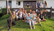 Eine Gruppe von Menschen sitzt auf einer Wiese vor einem Haus. © NDR Foto: Robert Tschuschke