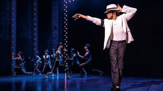 Ein Michael Jackson Darsteller tanzt in einer typischen Pose mit den Beinen überkreuzt, einer Hand am Hut und einem Arm nach vorn gestreckt. Es ist eine Szene aus "MJ - Das Michael Jackson Musical" auf der Bühne im Stage Theater an der Elbe in Hamburg. © Stage Entertainment Foto: Matthew Murphy
