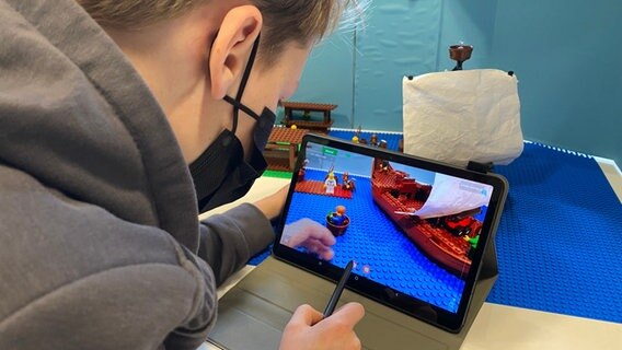 Der elf-jährige Finn rückt die Legofiguren auf blauem Untergrund in das rechte Licht. Dabei schaut er über ein Tablet auf das Geschehen und fotografiert die Szene. Der elf-jährige Finn rückt die Legofiguren auf blauem Untergrund in das rechte Licht. Dabei schaut er über ein Tablet auf das Geschehen und fotografiert die Szene. © NDR / Linda Ebener Foto: Linda Ebener
