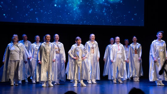 Kinder und Jugendliche mit silbernen Umhängen stehen auf einer Theaterbühne. © NDR Foto: Thomas Lorenzen