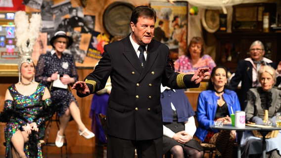 Ein Schauspieler in einer Pilotenuniform steht im Vordergrund und im Hintergrund sind andere Schauspieler mit verschiedenen Verkleidungen. Ein Schauspieler in einer Pilotenuniform steht im Vordergrund und im Hintergrund sind andere Schauspieler mit verschiedenen Verkleidungen. © NDR Foto: Michael Ermel