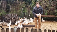 Jannik Rahlf, ein junger Mann mit brauner Hose und Anorak, steht mit Huhn auf dem Arm in einem Hühnerauslauf. © NDR Foto: Hannah Böhme