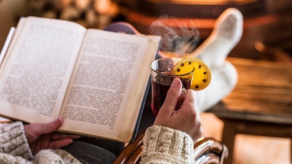 Eine Person sitzt in einem Sessel und liest ein Buch. In der rechten Hand wird ein roter Tee mit einer mit Nelken gespickten Orangenscheibe daran gehalten. Eine Person sitzt in einem Sessel und liest ein Buch. In der rechten Hand wird ein roter Tee mit einer mit Nelken gespickten Orangenscheibe daran gehalten. © fotolia Foto: volff