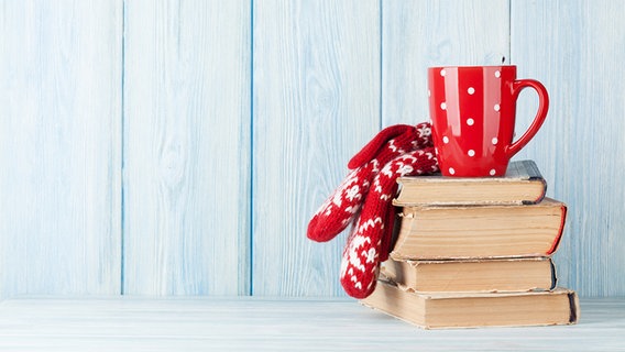 Auf einem Stapel Bücher steht eine rote Tasse mit weißen Punkten. Daneben liegen rot-weiße Handschuhe. © fotolia Foto: karandaev